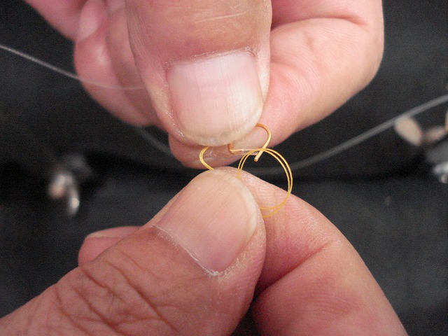 編込み台の片側のみ糸をはずす。はずした糸とホルダーにセットしてある糸とをあわせ持ち、手のひらと指（親指、人差し指以外）で押さえる。写真のような向きに左手でハナカンを持ち、右手で輪を作ってチューブの中に通す